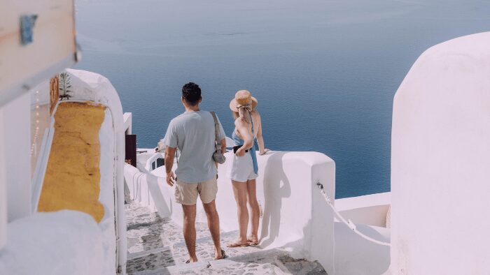 Couple standing on a whitewashed path in Santorini overlooking the Aegean Sea, searching for their transfer after arrival.