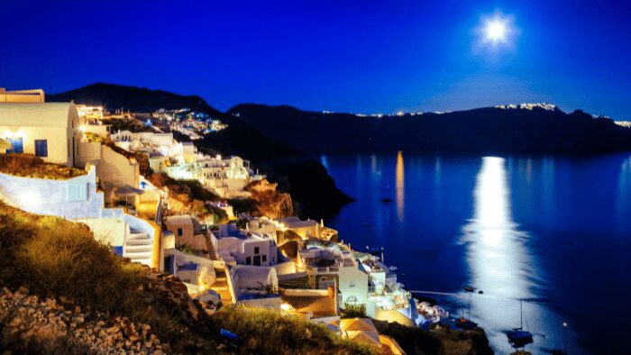 Night view of Oia village in Santorini with illuminated white houses overlooking the Caldera under a bright moon, capturing the island’s serene evening atmosphere.