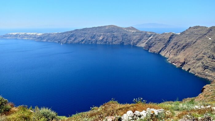 Panoramic view of the Santorini caldera from the Fira to Oia hiking trail, with deep blue water and white village visible on the clifftop in the distance