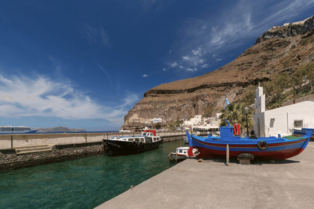 Small Old Port harbour at the base of volcanic caldera cliffs. A colourful blue and red fishing boat is docked in the foreground. Tender and excursion boats are moored along the quay. Whitewashed buildings and the cable car infrastructure are visible on the cliff face. A large cruise ship is anchored in the caldera in the background left. Clear blue sky.