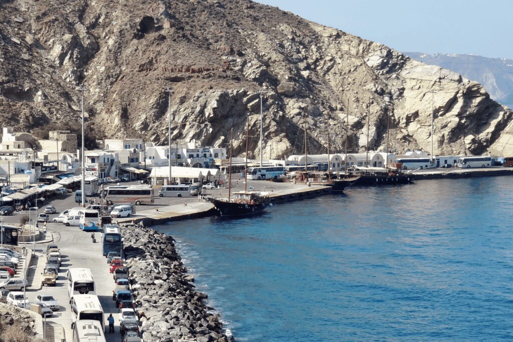 Busy road-accessible port Athinios in Santorini. Multiple coaches, minivans, cars, and buses line the harbour road. A tall-masted sailing vessel is docked at the pier. White commercial buildings and storage structures run along the waterfront. Volcanic rock cliff backdrop. Clearly a working transport hub with heavy vehicle activity.