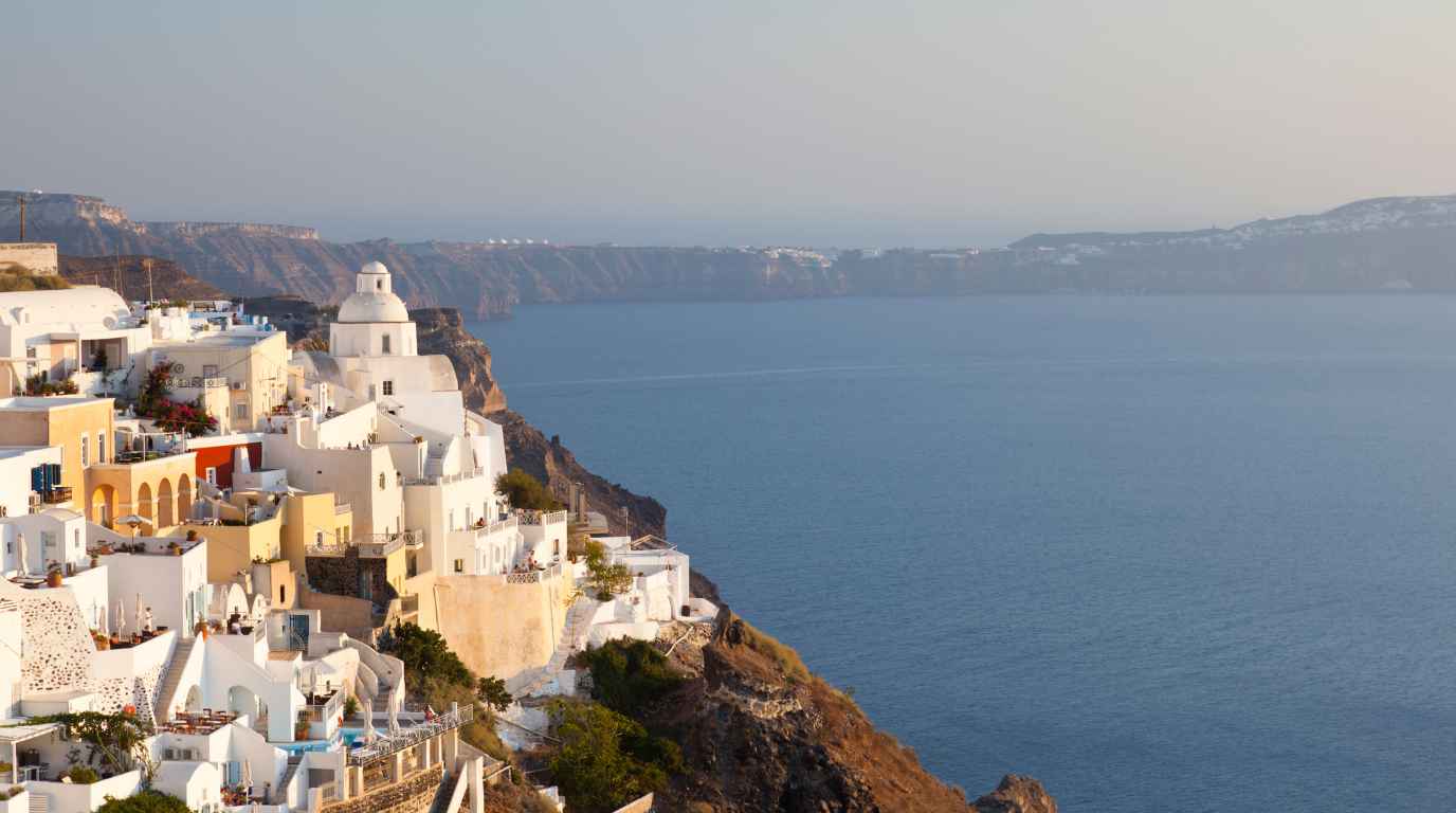 Whitewashed clifftop houses and a domed church on the Santorini caldera rim above the deep blue Aegean at golden hour
