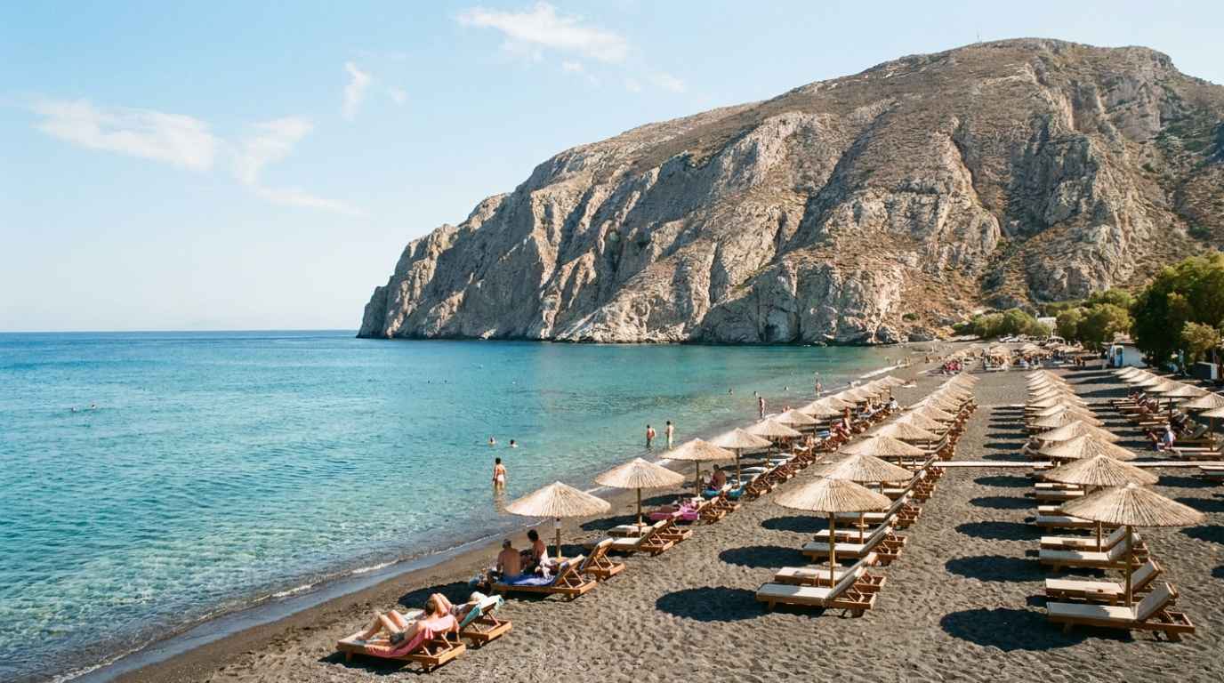Black sand beach at Kamari, Santorini, with sun loungers and the Mesa Vouno headland